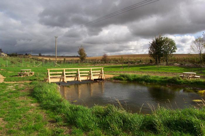 Pond with wooden bridge in nature area with picnic tables, grass, trees and cloudy sky